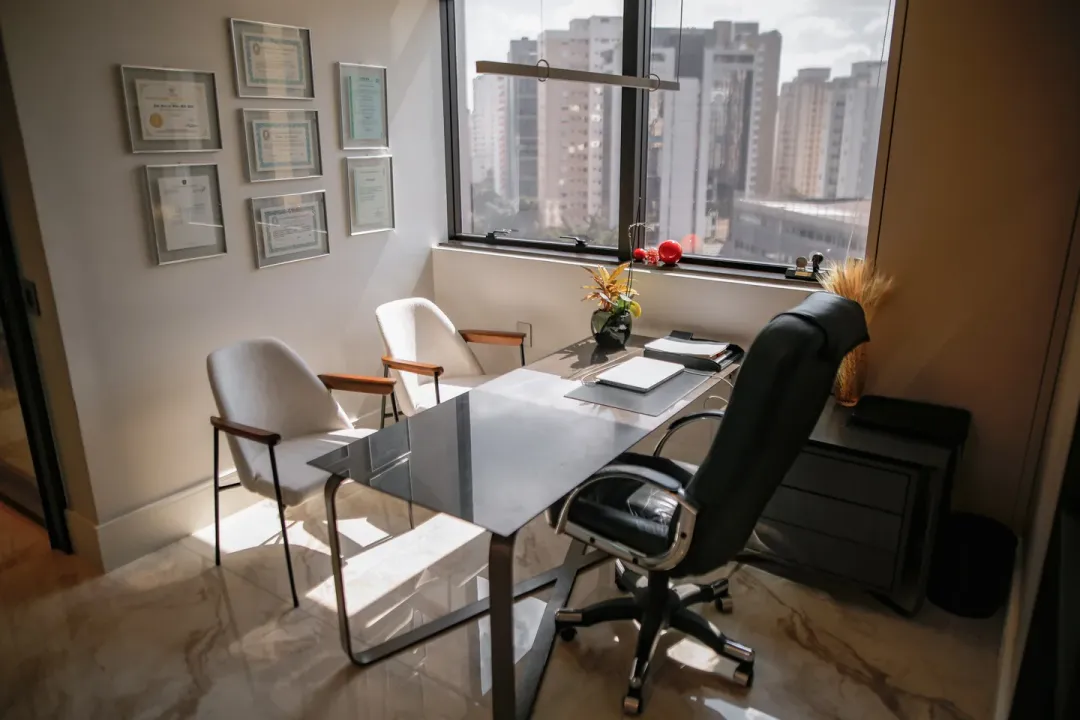 Sala de consulta vista de outro ângulo, com as cadeiras do paciente, os diplomas na parede e a vista da cidade pela janela